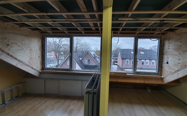 Het interieur van een zolder met een nieuwe prefab dakkapel, met zicht op omliggende huizen door de ramen.